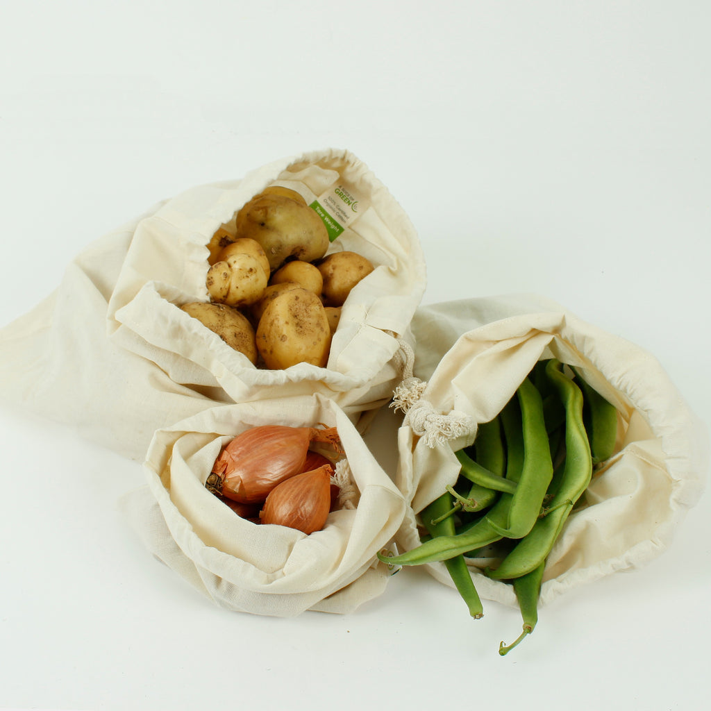 Three cream drawstring cotton produce bags filled with potatoes, shallots and green beans on white background