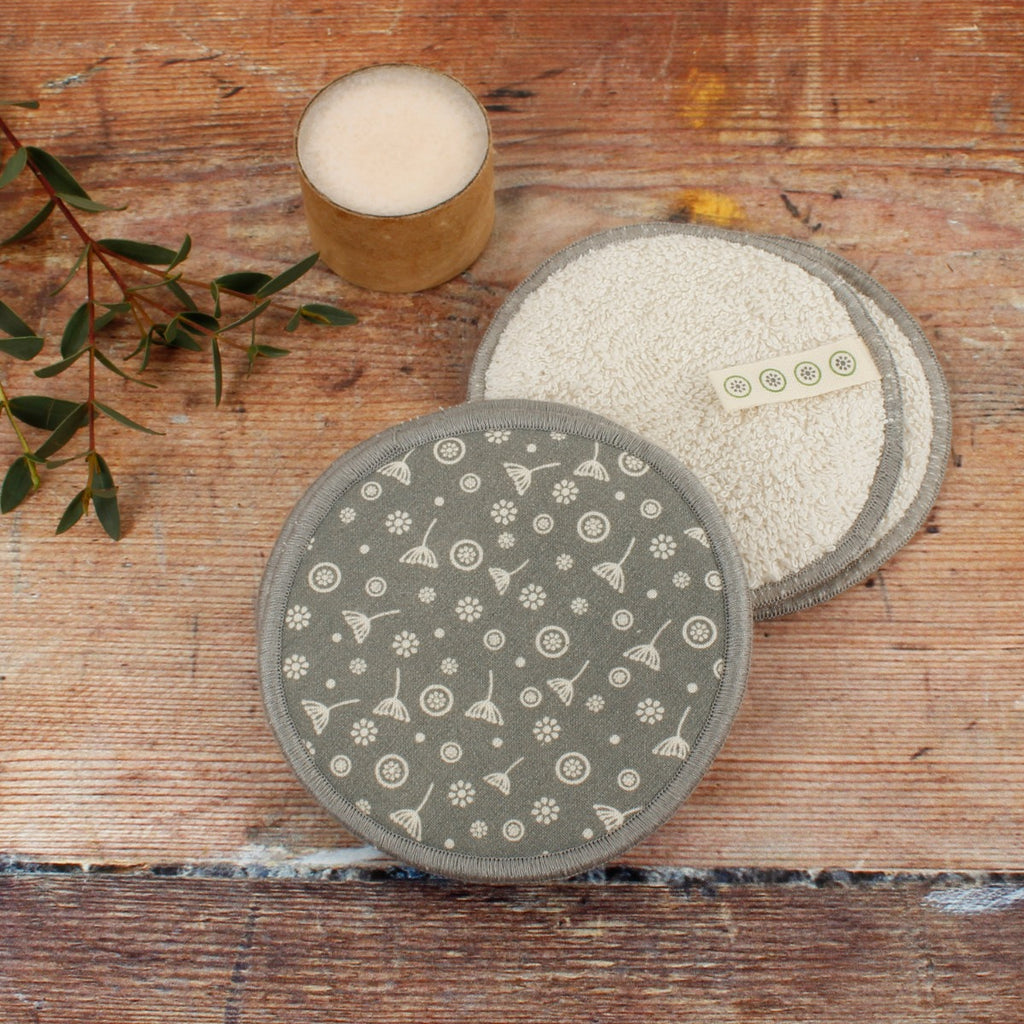 Organic cotton facial pads with grey floral print and soft white side on wood surface beside soap and greenery