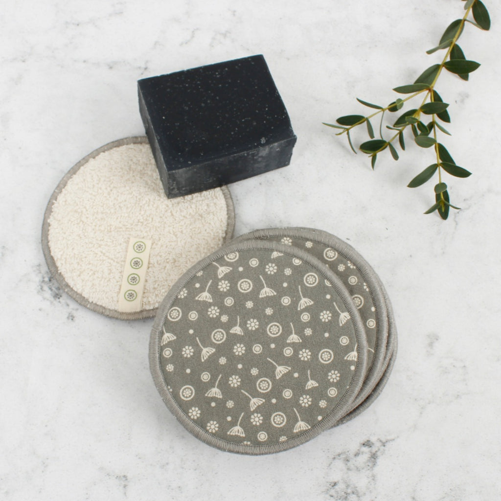 Reusable cotton facial pads with grey floral print and soft side on marble surface beside black soap and greenery