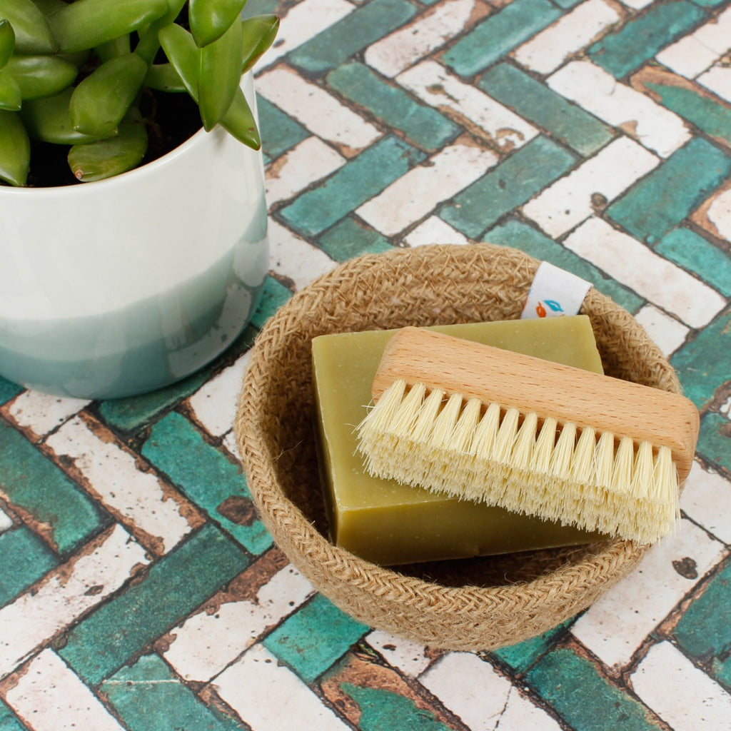 Wooden nail brush and green soap bar in a jute dish beside a potted plant on a patterned tile surface