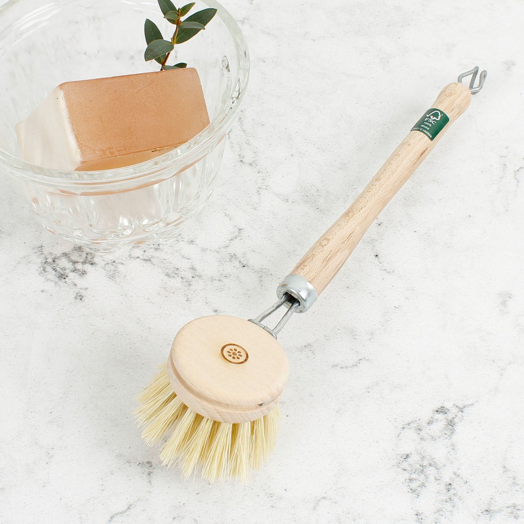 Wooden dish brush with long handle and round head on marble surface beside clear bowl with soap and greenery