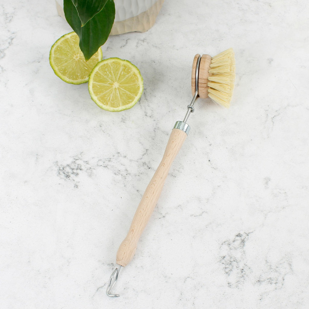 Wooden scrub brush with metal hook and round bristle head on marble surface beside sliced lime and green foliage