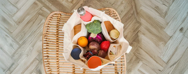 A canvas tote bag filled with fresh groceries like broccoli, oranges, and jars, placed on a woven chair over a herringbone wood floor.