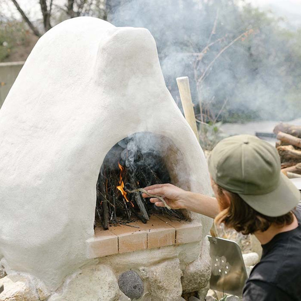 Person lighting kindling in an outdoor clay oven using a firestarter, with smoke rising from the small flame