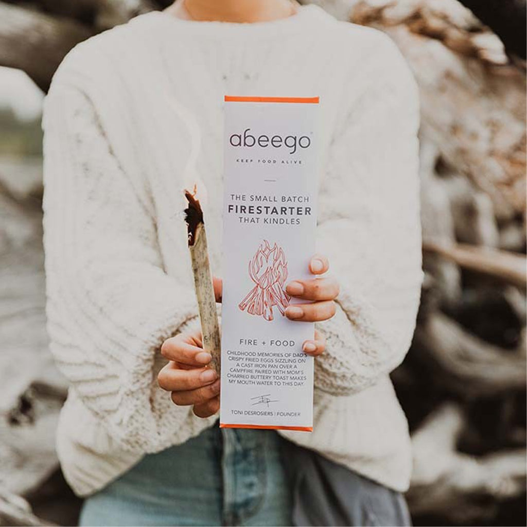 Person in a cream jumper holding an Abeego firestarter pack and a smoking firestarter stick outdoors