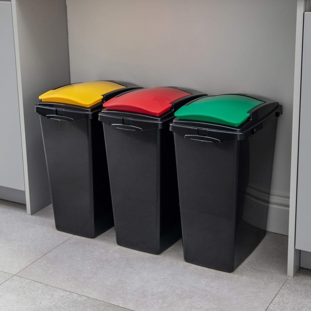 Set of three black recycling bins with yellow, red and green lids, neatly placed under a kitchen counter for organised waste separation.