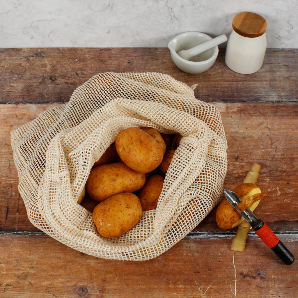 Organic cotton mesh produce bag filled with potatoes on wooden surface beside peeler, skins, and kitchenware