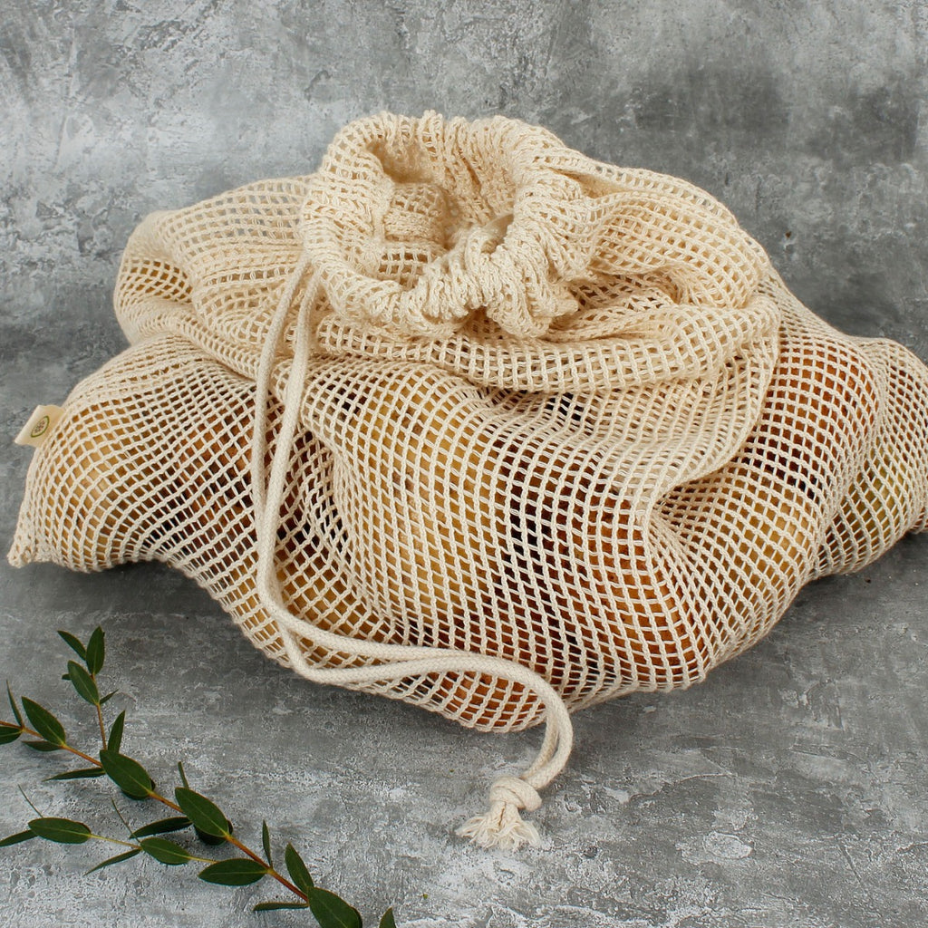 Drawstring organic cotton mesh bag filled with potatoes on grey surface beside a sprig of green leaves