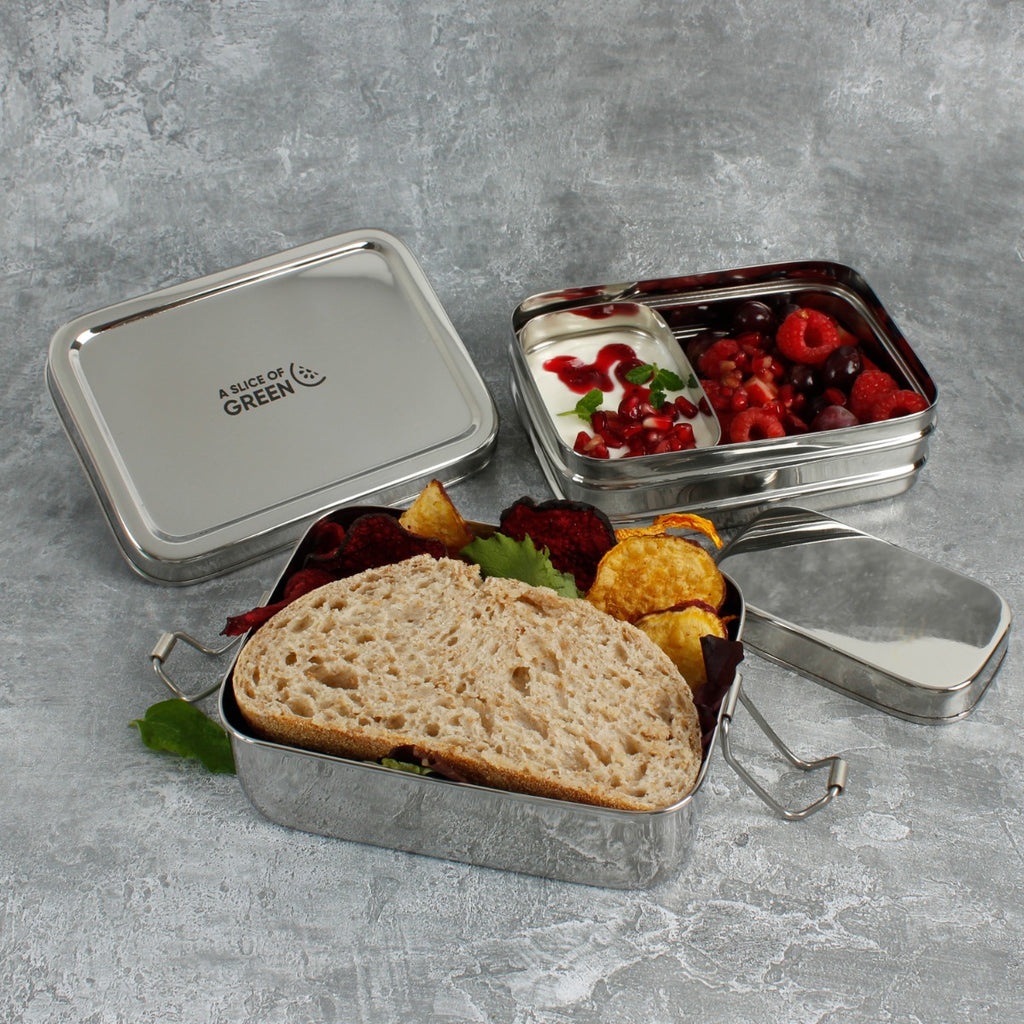 Two-tier stainless steel lunch box open with bread, salad, fruit, and yoghurt inside, displayed on a textured grey surface