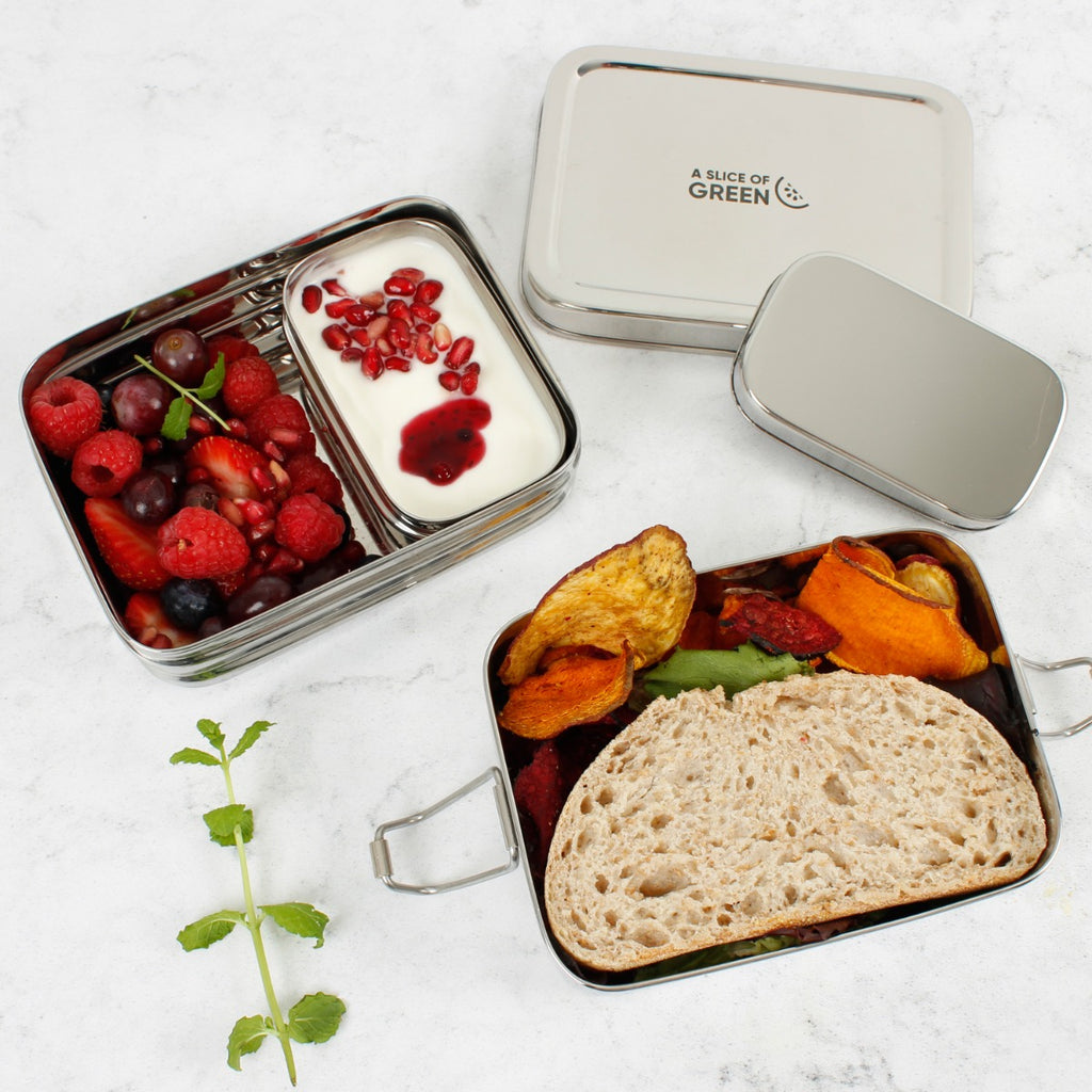 Two-layer stainless steel lunch box open with bread, veggie crisps, fruit, and yoghurt on a white background with mint garnish