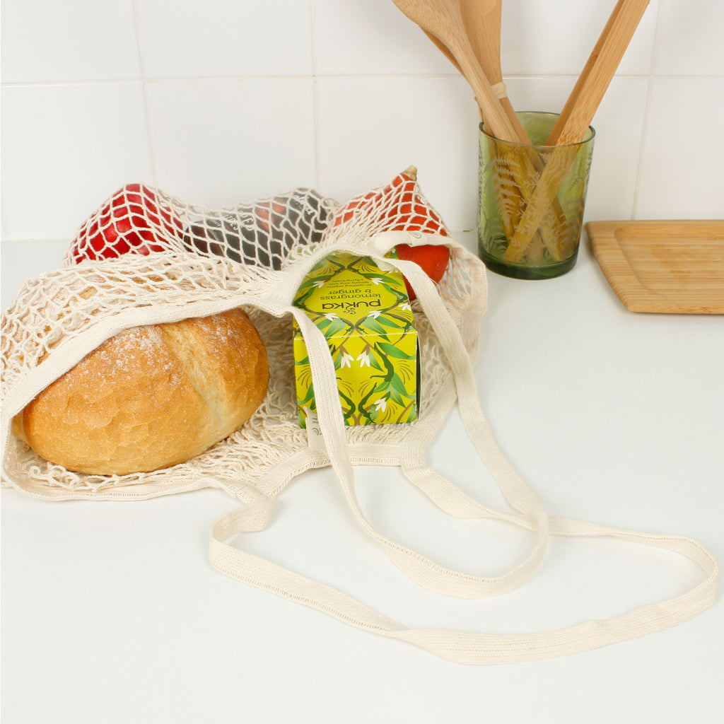 Organic cotton mesh shopping bag on kitchen counter filled with bread, fruit, and herbal tea, long handles spread out in front