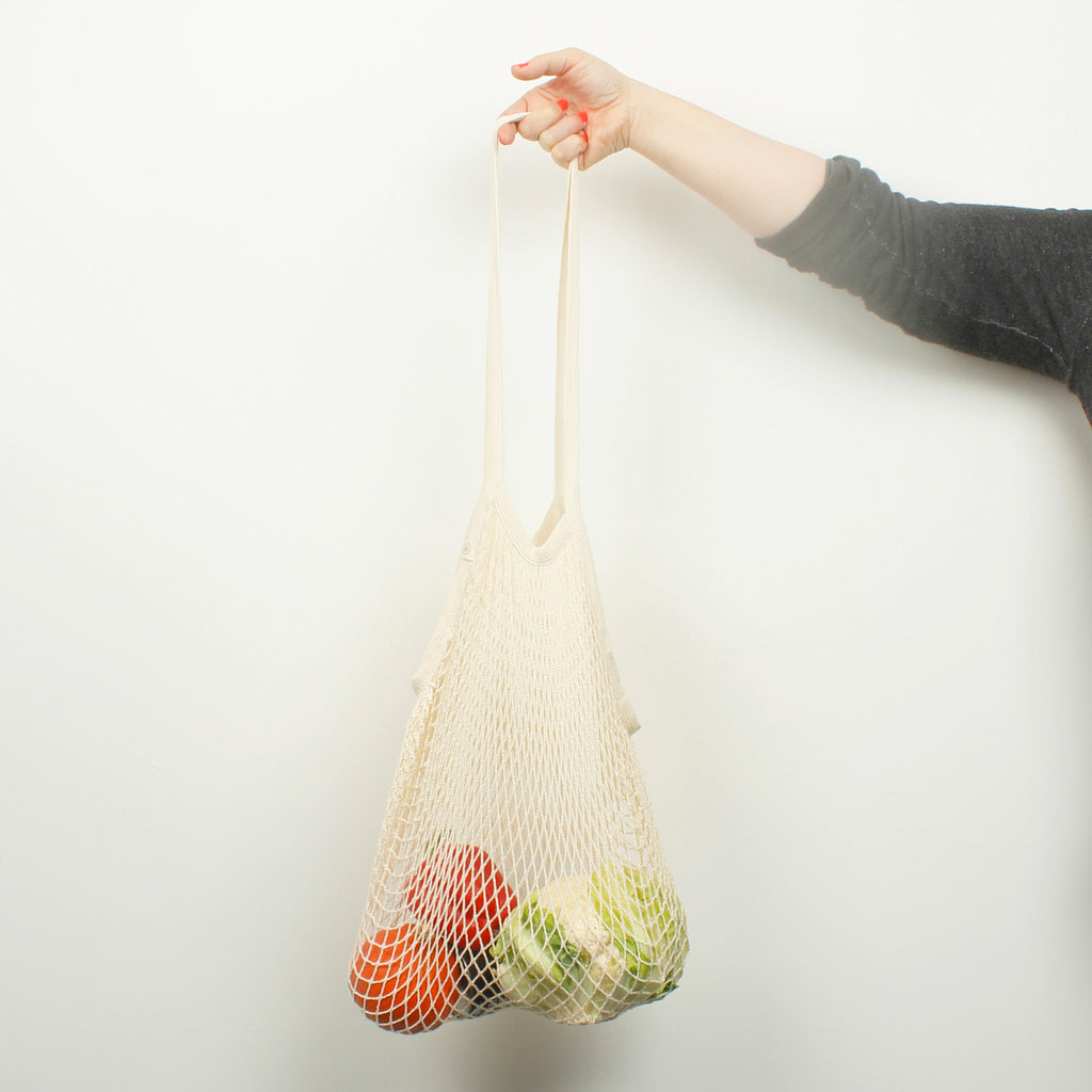 Arm holding organic cotton mesh shopping bag filled with fresh produce against plain white background