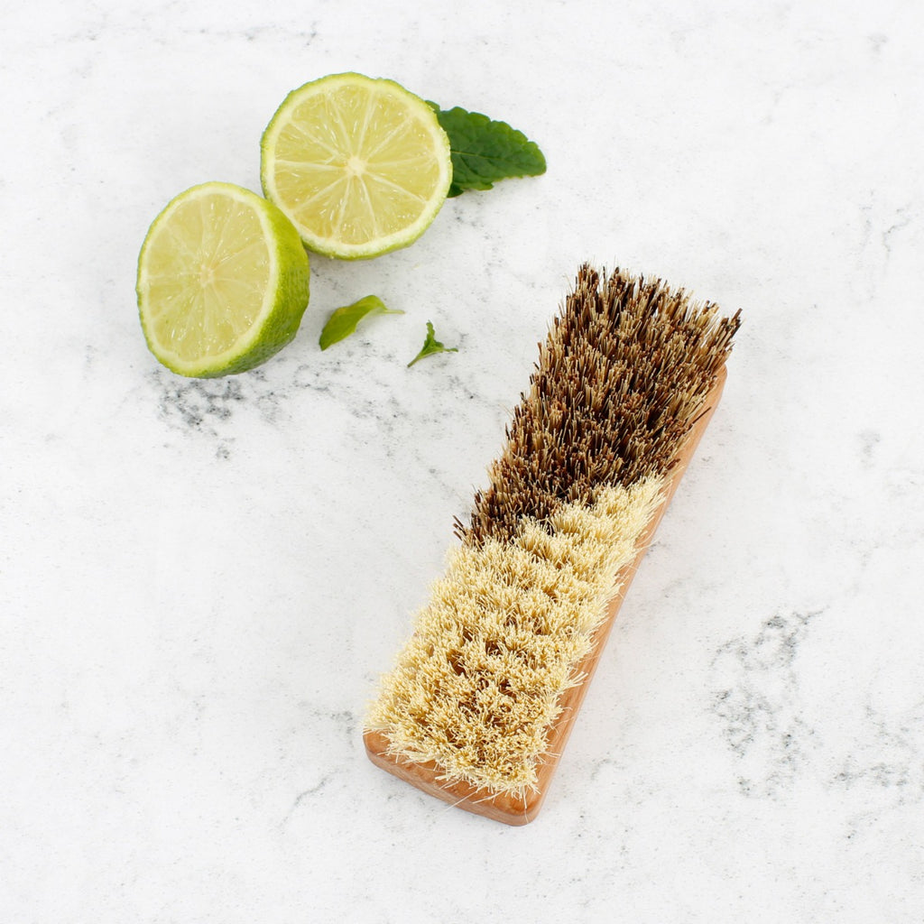 Wooden vegetable brush with dual bristles on marble surface beside lime halves and mint leaves